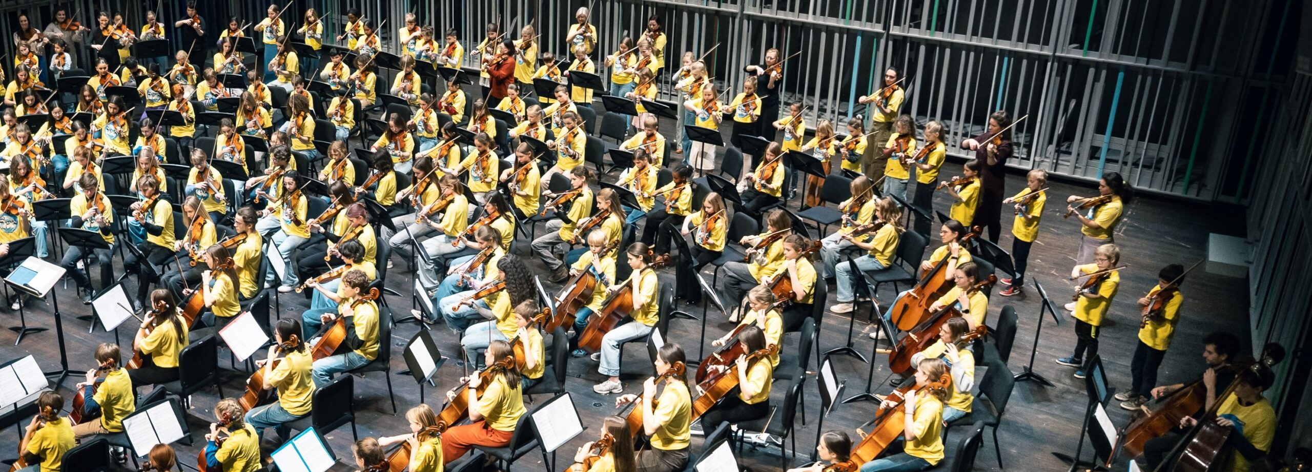 Een 100-tal kinderen zit in een halve cirkel als orkest op een podium. Ze dragen allemaal een gele t-shirt en spelen een strijkinstrument.